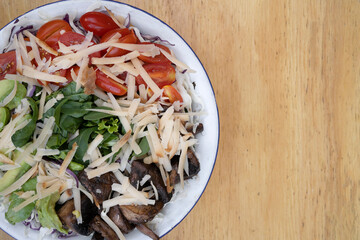 Top view of a healthy salad with cherry tomatoes, mushrooms, avocado, spinach, arugula and provolone cheese, in a white bowl on the wooden table.