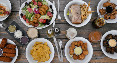 Panorama view of the restaurant wooden table with delicious dishes and drinks such as salad, nachos, fried shrimps, spinach fritters, sandwich, fried chicken wings, bread, mozzarella sticks and beer