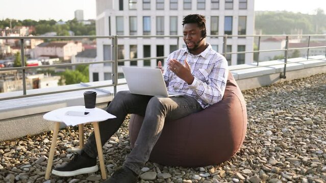 Relaxed African American man conducting web conference via laptop and headset while staying on flat roof. Professional entrepreneur working outside of corporate offices via wireless devices.