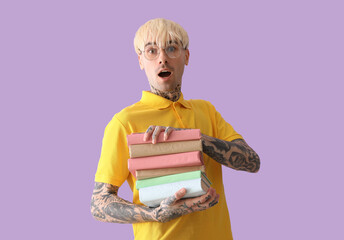 Shocked tattooed young man with stack of books on lilac background