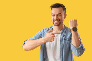 Handsome young man pointing at wristwatch on yellow background