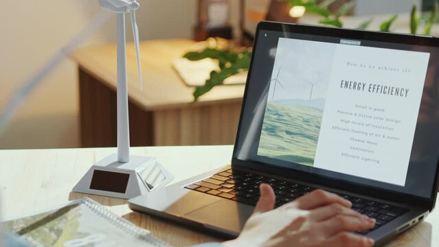 Hands of unrecognizable female engineer sitting at office desk with wind turbine model and looking at presentation slides on laptop. Close-up view