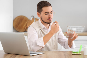 Young man with laptop and glass of water taking pills at home