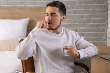 Young man with glass of water taking pills in bedroom