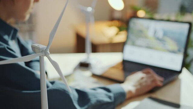 Close-up view of wind turbine model in the office, female green energy engineer using laptop at workplace in defocused background