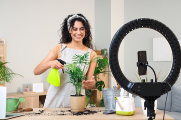 Obraz premium Female African-American gardener with plant and spray water bottle recording video at home