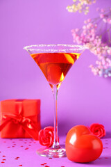 Glass of tasty cocktail with hearts, roses and gift on purple background, closeup. Valentine's Day celebration