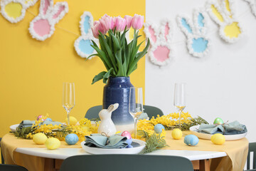 Festive table setting decorated with mimosa flowers, vase of tulips, Easter bunny and painted eggs