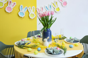 Festive table setting decorated with mimosa flowers, vase of tulips, Easter bunny and painted eggs