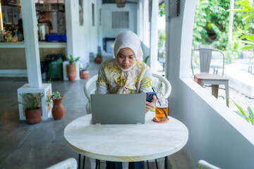 A beautiful, charismatic Asian woman wearing a casual hijab is working or doing assignments in a cafe. Seen using a laptop. The concept of working anywhere. - obrazy, fototapety, plakaty