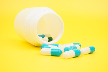Scattered white and blue pill capsules spilled out of a white bottle isolated on yellow background
