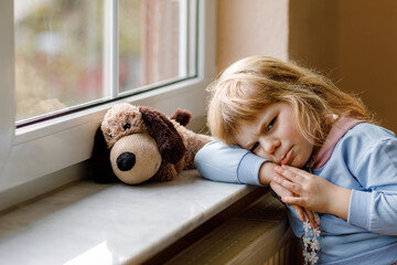 Upset toddler girl sitting by window and looking out. Sad crying child feeling unhappy. Lockdown...