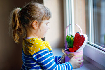Little girl sitting by window with hyacinth flowers and hand-made heart. Happy child, indoors....