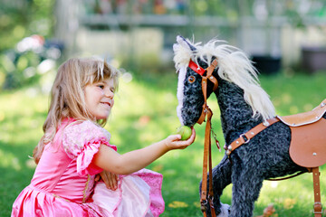 Little preschool girl hugging with rocking horse toy. Happy child in princess dress on sunny summer day in garden. Girl in love with her favourite old vintage toy animal.