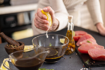 Woman squeezes fresh lemon juice into glass bowl with soy sauce and olive oil making marinade to...