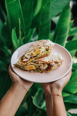 A person's hand is holding a plate filled with burritos with cheese, vegetables, and meat, and behind is greenery
