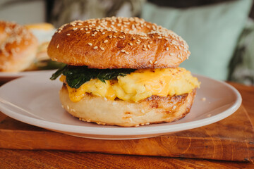 A close-up of a bagel burger with scrambled eggs, spinach, cheese, and avocado on a plate