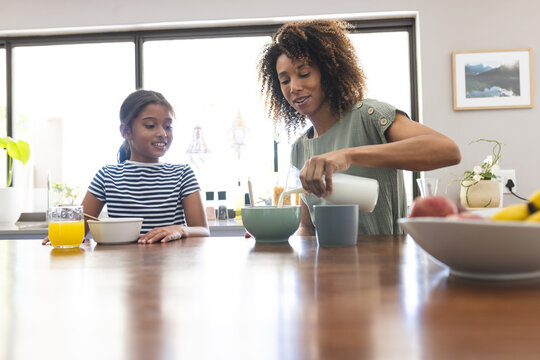 Biracial mother pours milk and cereal at home for her daughter, with copy space