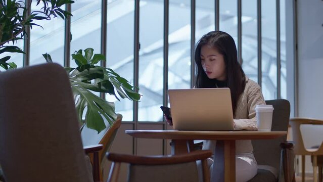 Pretty Asian girl works on  laptop in cafe, checking messages on her phone. Open shot.