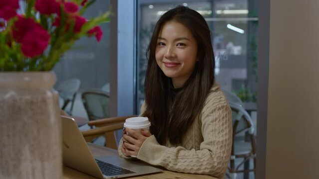 Young pretty Asian woman smiles on camera in cafe. Dolly in shot.