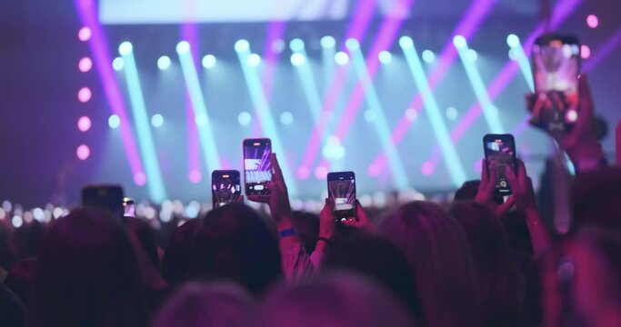 Fans film popular music band concert on phones, close up. Viewers crowd record bright neon light show for social media. Lot fun people enthusiastically take photos of music festival on smartphones