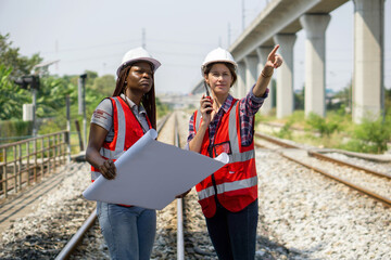 Two construction or engineer with safety helmet and high-visibility vest reviewing work to be done or progress on a railway track project while holding a set of plans or blueprint and walkie-talkie.