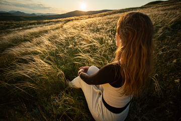 A young adult woman in a yoga suit sits in the green grass of feather grass and enjoys the warm...