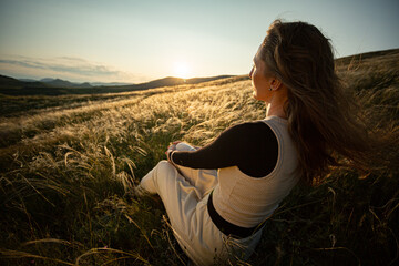 A adult woman sits in the green grass of feather grass and enjoys the warm rays of the sunset with...