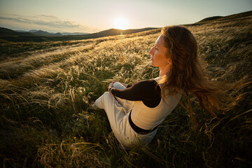 A young adult woman sits in the green grass of golden feather grass and enjoys the warm rays of the...