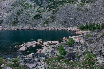 Summer nature landscape. Secluded shore of a pond. Emerald colored mountain lake surrounded by stones and evergreen coniferous trees. 