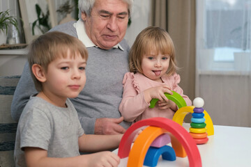 Grandfather playing toys with grandson and young granddaughter indoor. Old man grandpa studying...