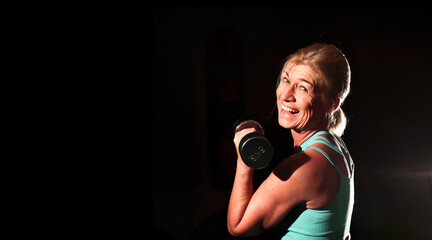 Woman in her mid-fifties training in the gym, photographed with flash against a black background.