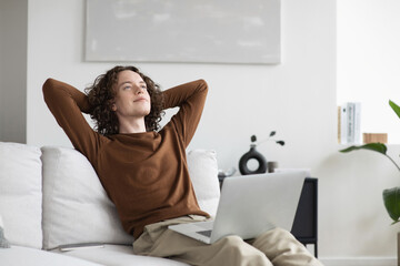 Satisfied handsome young man relaxing on sofa at home in living room, resting after a hard day work, put hands behind head, smiles happily. Relaxation, self care, enjoy life concept