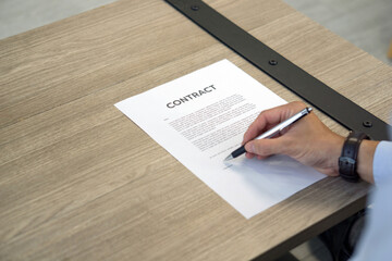 Closeup CONTRACT document on wooden table, man hand with wristwatch holding a pen signing on it.
