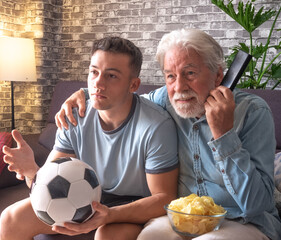 Caucasian couple of young boy and senior grandfather soccer fans watching a football game on tv sitting on a comfortable sofa in living room