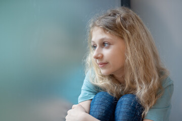 Thoughtful girl sitting on sill embracing knees looking at window, sad depressed teenager spending time alone at home