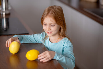 Smiling beautiful girl holding yellow lemons standing  in kitchen at home. Portrait of pretty child choosing food of healthy products