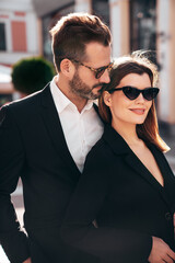 Beautiful fashion woman and her handsome elegant boyfriend in black suit. Sexy brunette model in jacket. Fashionable confident couple posing in street. Brutal man and female outdoors. In sunglasses