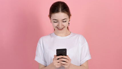 Fototapeta premium Attractive Caucasian young brunette woman in casual white t-shirt smiling and using mobile phone isolated on pink studio background. Freelance, network, online work.
