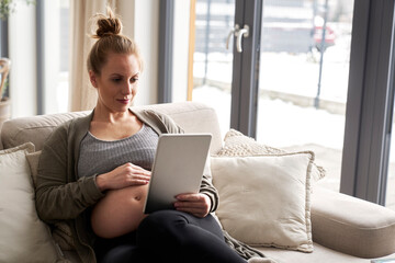 Caucasian pregnant woman sitting on sofa and using digital tablet