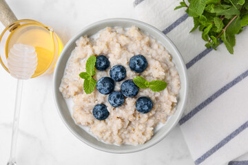 Delicious barley porridge with blueberries and mint served with honey on white marble table, flat lay