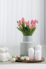 Easter decorations. Bouquet of tulips, painted eggs, burning candles and bunny figures on white table indoors