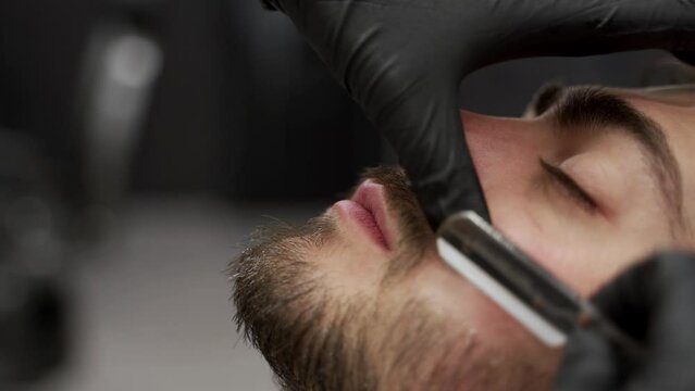 Professional barber shaves clients beard in upscale barbershop. Male pampering with straight razor, expert grooming skills in luxurious salon. Gentlemans beard care, precise facial hair styling.