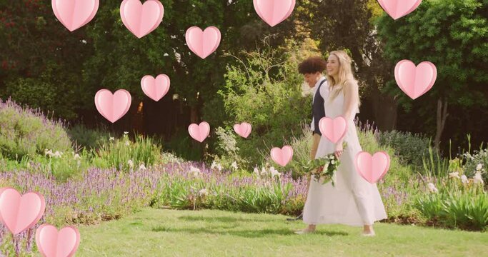 Animation of pink hearts over happy diverse couple walking in sunny garden on wedding day