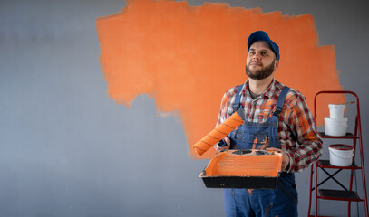Painter man holding a paint roller and plastic tray on orange wall background thinking an idea...