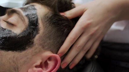 Barber applies black charcoal mask on clients face in shop. Man relaxes during skincare routine. Grooming ritual, pore cleansing, detox treatment. Male pampering for clear skin, facial care.