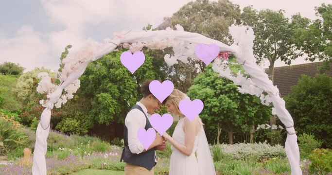 Animation of lilac hearts and happy diverse couple holding hands under wedding arch in garden