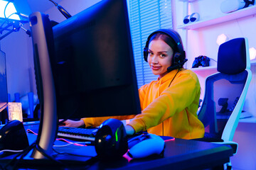 Young Caucasian woman professional gamer wearing yellow hoodie sits on a chair with a gaming table...