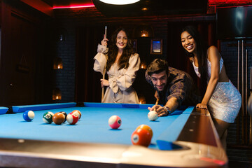 Young diversity group of people playing pool together with smile, enjoyment and fun. Young people spend time in billiards room at the nightclub. Men, women friends playing billiards. Nightlife concept - obrazy, fototapety, plakaty