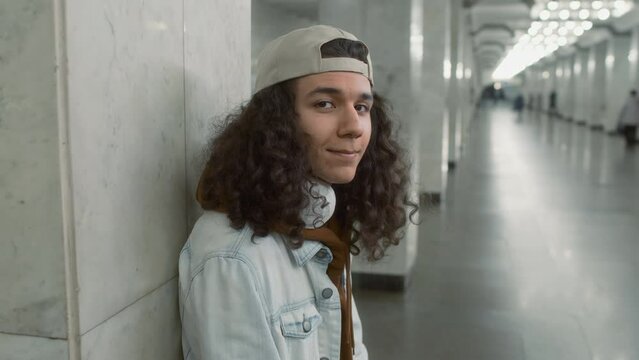 Push in side portrait of Caucasian positive curly-haired young man in cap and with headset on neck looking at camera while posing in modern light metro station and leaning on tiled pillar